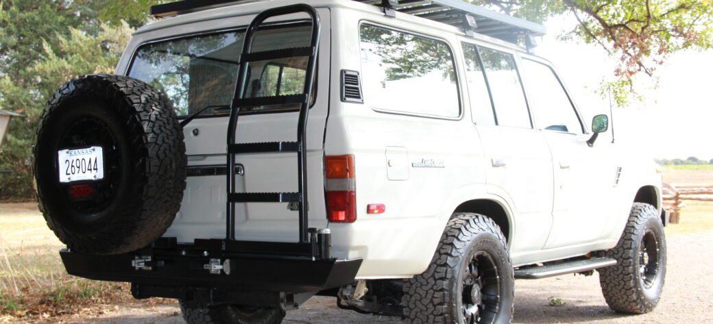 1990 FJ62 Toyota Land Cruiser V8 conversion in white, rear three-quarter view with roof rack, rear ladder, custom bumper with tire carrier, and BFG KO2 tires