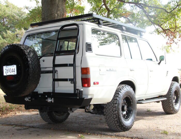 1990 FJ62 Toyota Land Cruiser V8 conversion in white, rear three-quarter view with roof rack, rear ladder, custom bumper with tire carrier, and BFG KO2 tires
